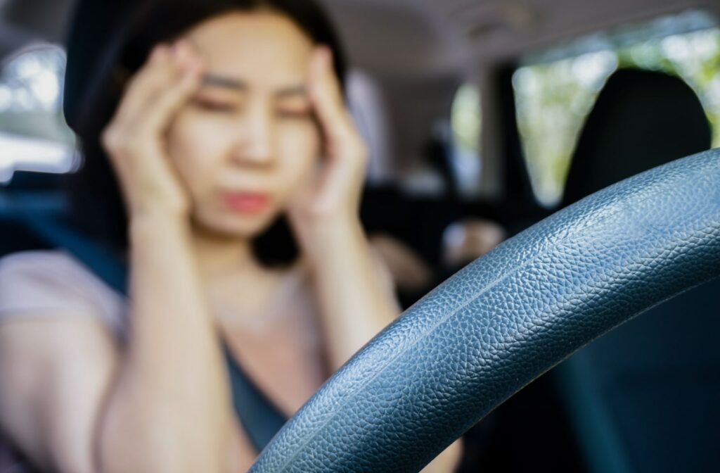 A woman sitting behind the wheel of a car, and she is holding her hands up to the temples of her head due to dizziness