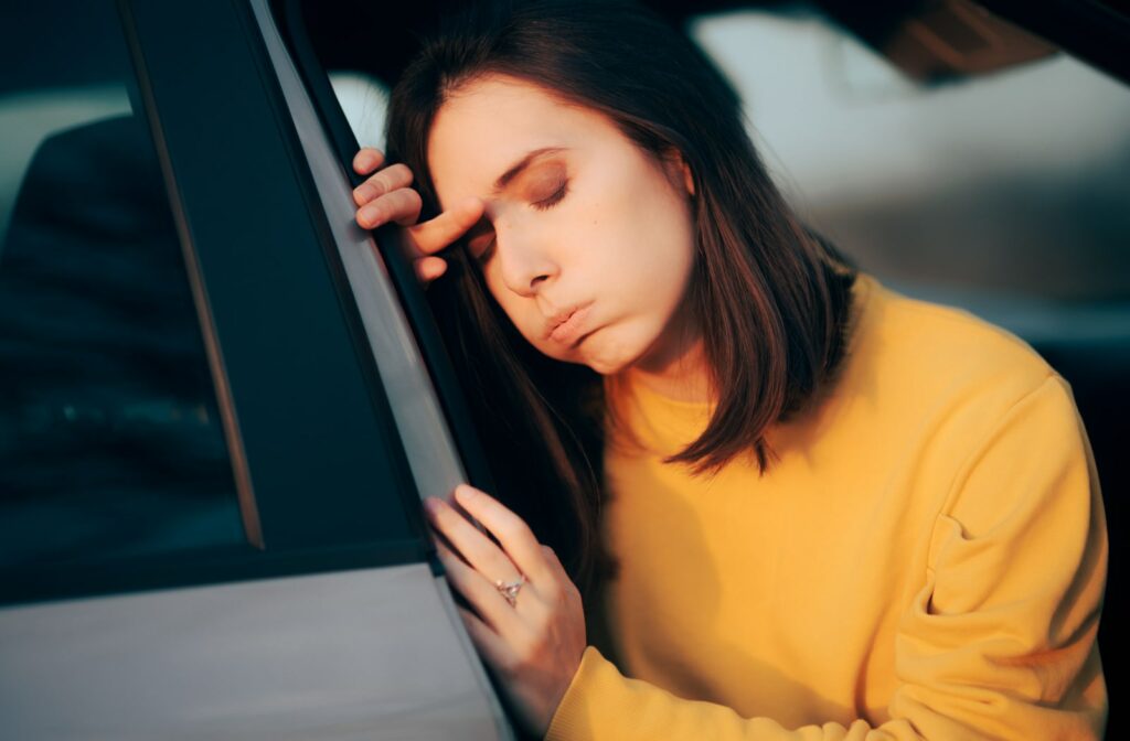 A woman wearing a yellow sweater in a car, and she's pulled off to the side of the road. She's sitting on the side of her seat by the car door and feeling unwell