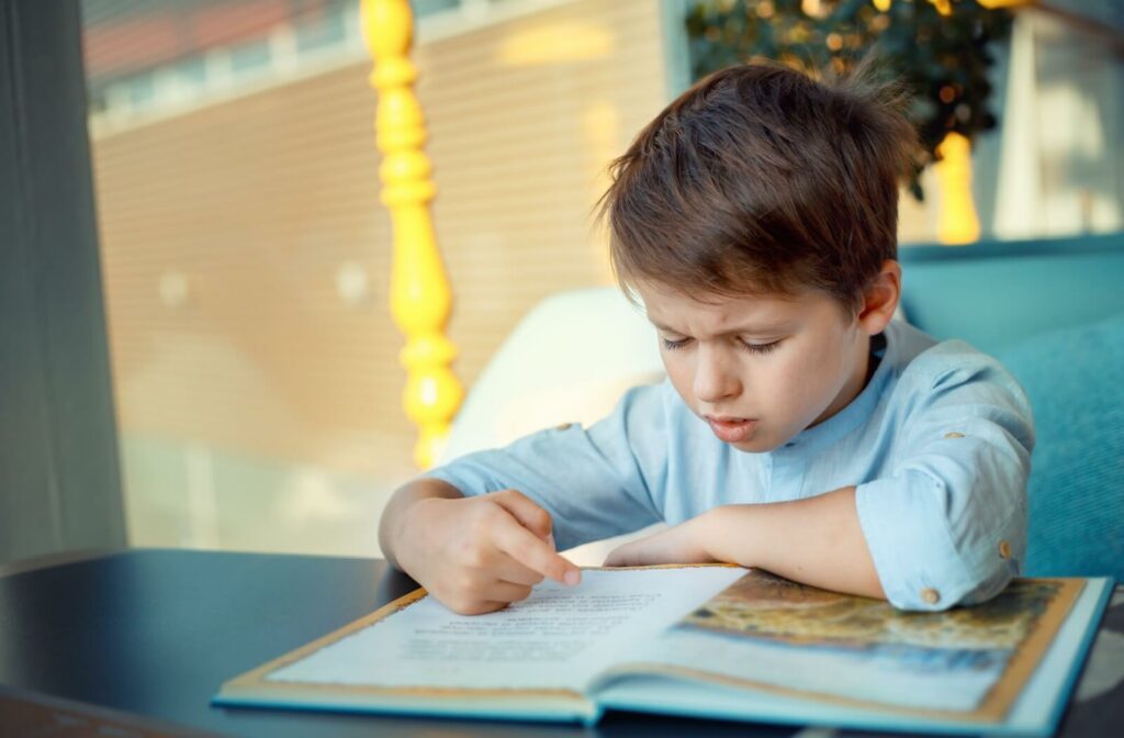 A young child points to words in a book with his right hand as he struggles to read.