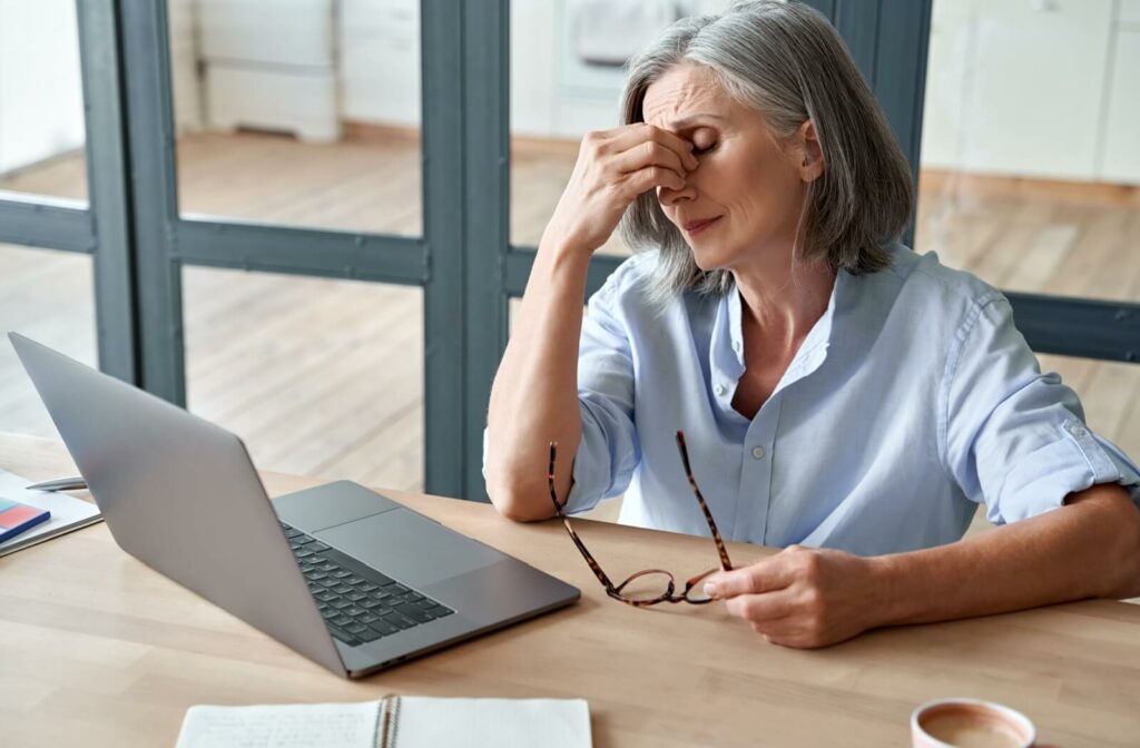 A woman working on a laptop removes her glasses and rubs her irritated and dry eyes