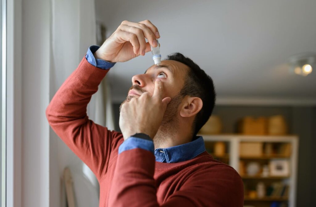 A person stands in their living room, holding one eye open while using the other hand to apply eye drops.