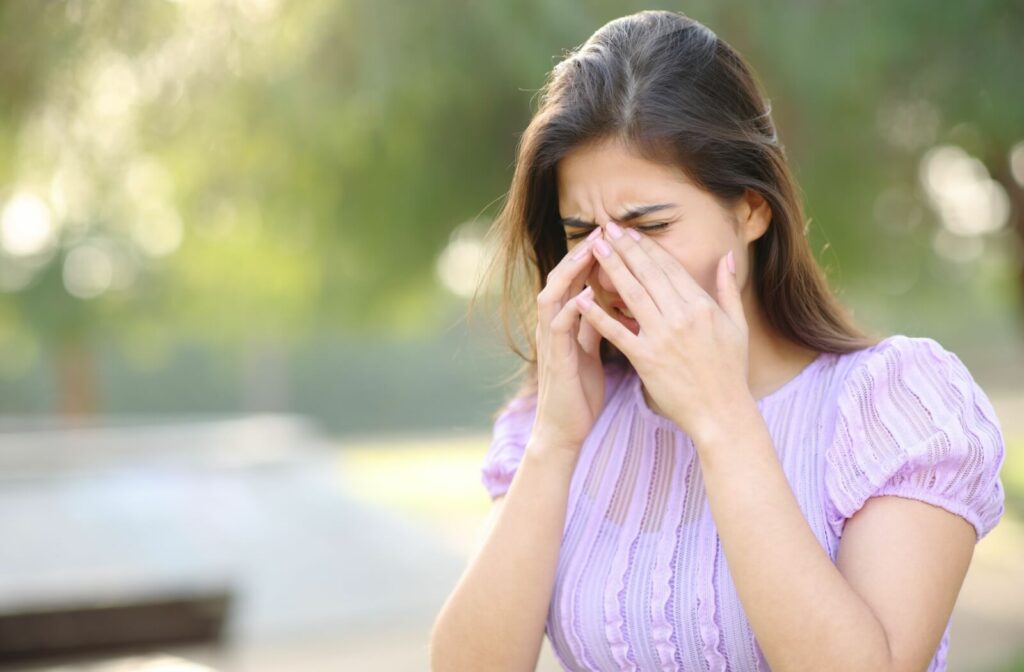 A person rubs their eyes while standing outside on a bright, sunny day. Trees are visible in the background, suggesting the person is at a park and experiencing seasonal allergies.