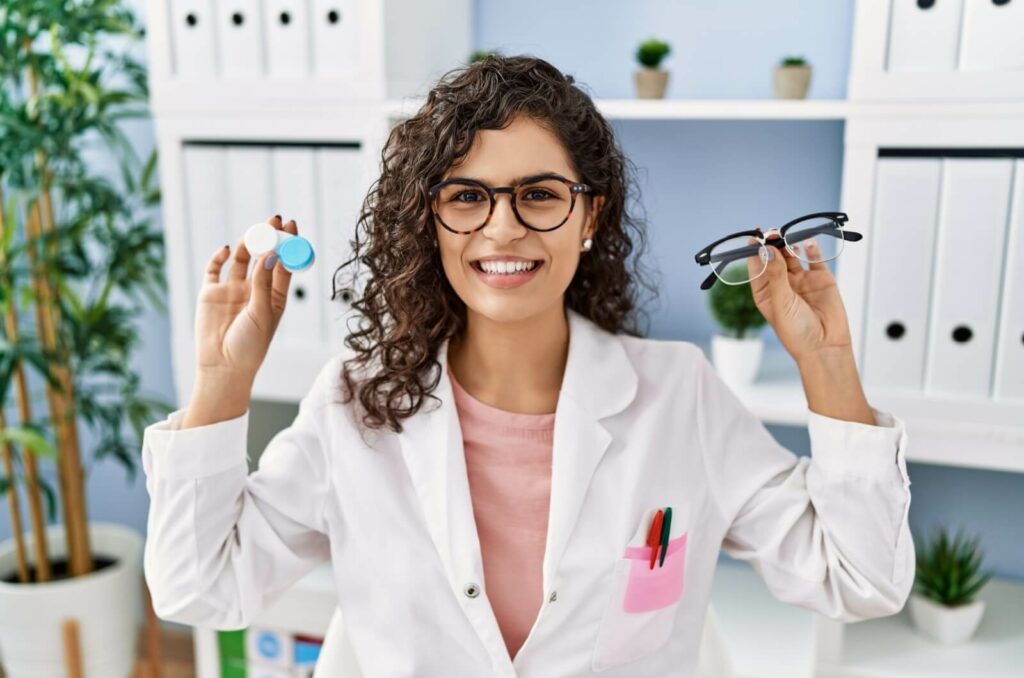 A young, happy optometrist holding a contact lens case in one hand and a pair of glasses in the other.