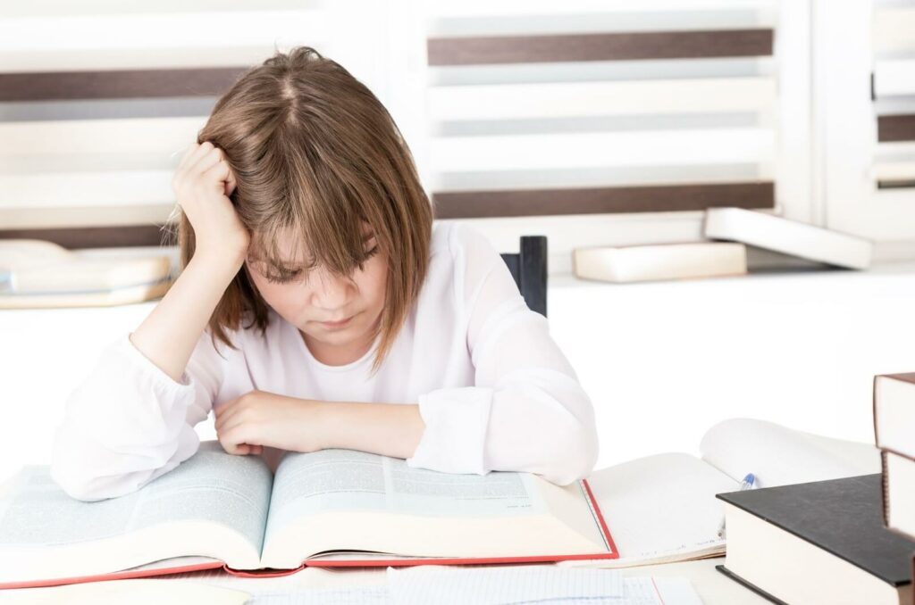 A frowning child holds their head with one hand while they struggle to read the book in front of them.