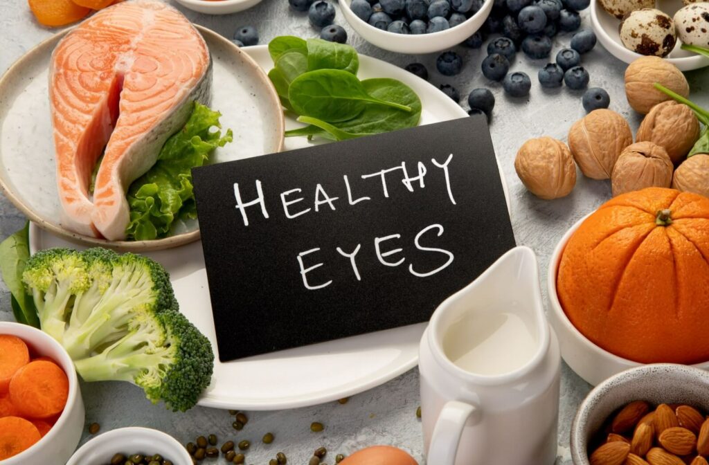 A black sign that reads “Healthy Eyes” sits among bowls of eye-healthy foods, including salmon, broccoli, and citrus fruits