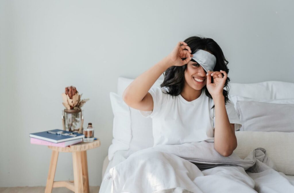 Person sitting in bed smiling while adjusting a gray eye mask suggesting comfort and relief for dry eyes