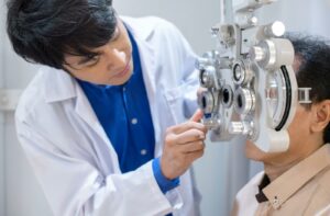 An optometrist in a white lab coat and blue collared shirt giving an eye exam to a patient in a bright clinical setting