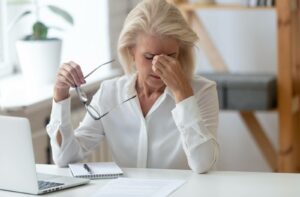 Older woman rubbing her eyes in discomfort while holding glasses, possibly experiencing blurry vision.