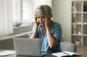 Older woman sitting at a desk, touching her temples with eyes closed, appearing to experience eye strain or vision discomfort while working on a laptop.