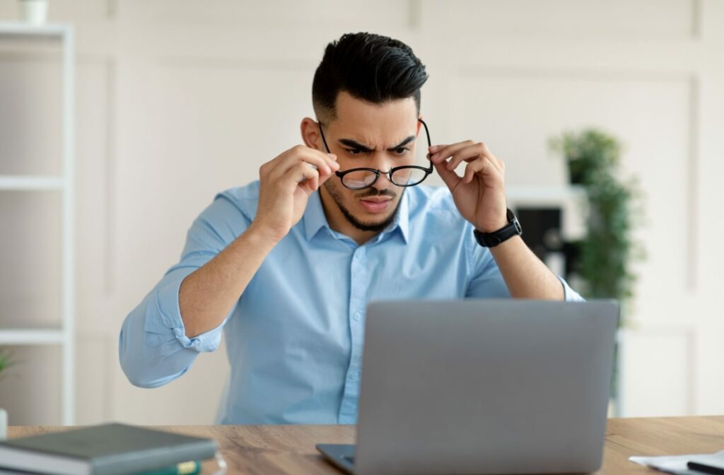 Man in a blue shirt squinting at a laptop screen while adjusting his glasses experiencing sudden blurry vision.