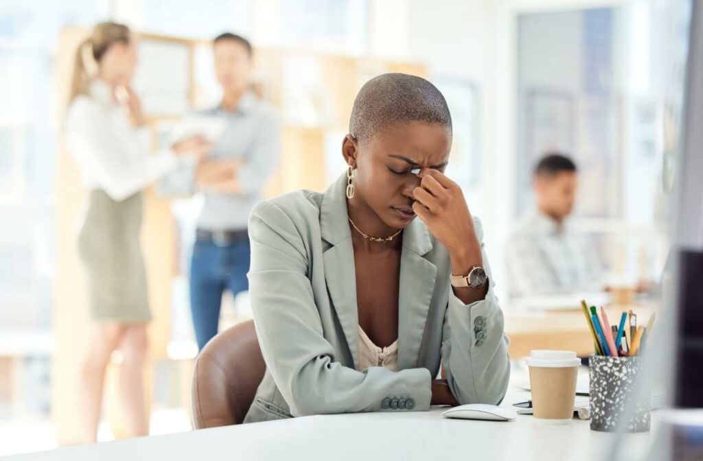 Woman with a headache sitting at her desk, holding the bridge of her nose with her eyes closed.