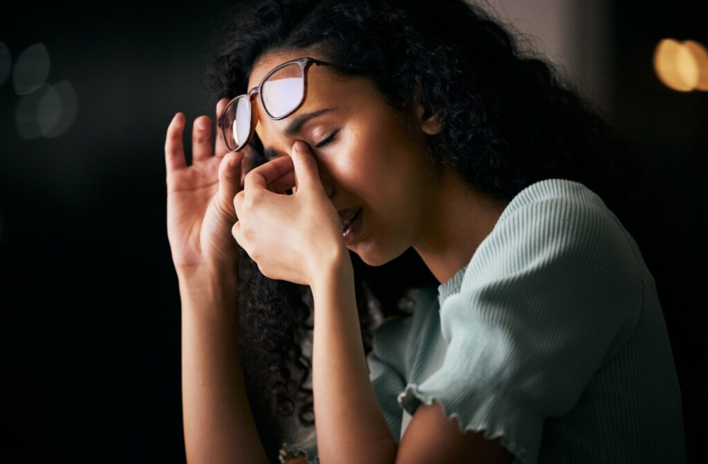 A person with curly hair rubbing her eyes in discomfort while holding her glasses above her forehead, suggesting eye strain or tiredness after prolonged screen time or focus.