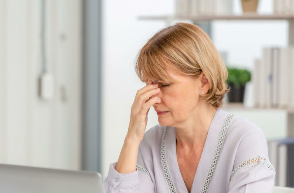 A person sitting at a desk with eyes closed, pressing their fingers to the bridge of their nose as if experiencing eye strain or fatigue.
