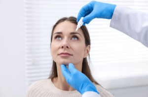 A person looking upward while an eye care professional wearing blue gloves applies eye drops during an examination.
