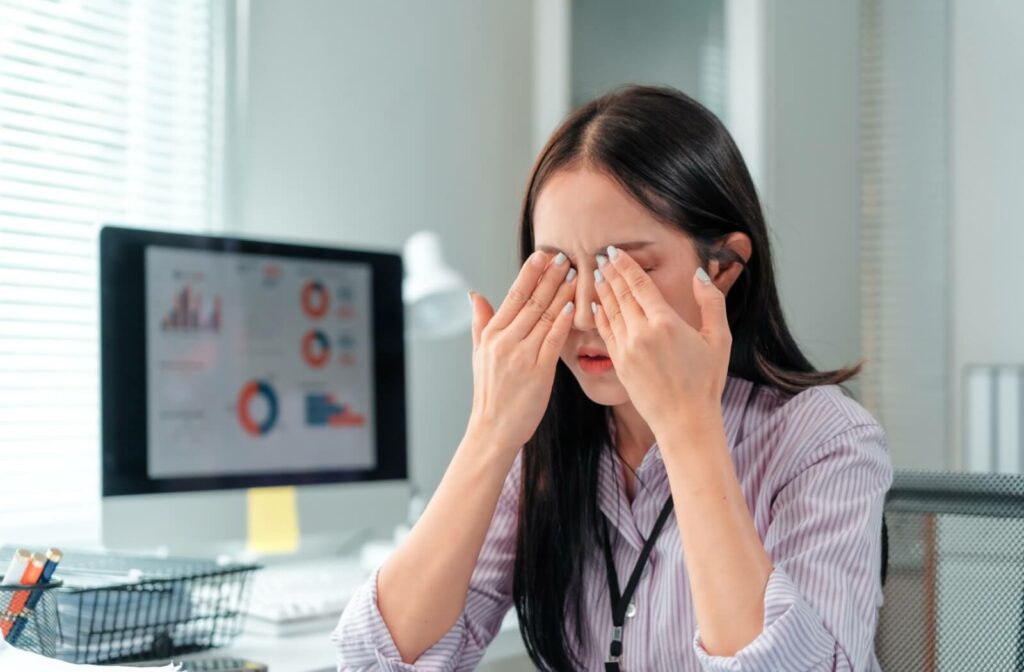 Woman experiencing eye strain and rubbing her eyes at her desk in front of a computer screen.