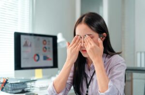 Woman experiencing eye strain and rubbing her eyes at her desk in front of a computer screen.