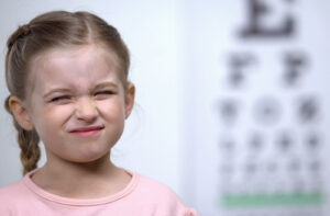 A young girl squinting and scrunching her face in front of a blurred eye chart, illustrating visual strain and difficulty focusing caused by eye misalignment.