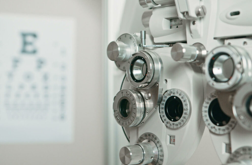 A close-up of a phoropter used for vision testing in an optometry clinic with a blurred Snellen eye chart in the background.