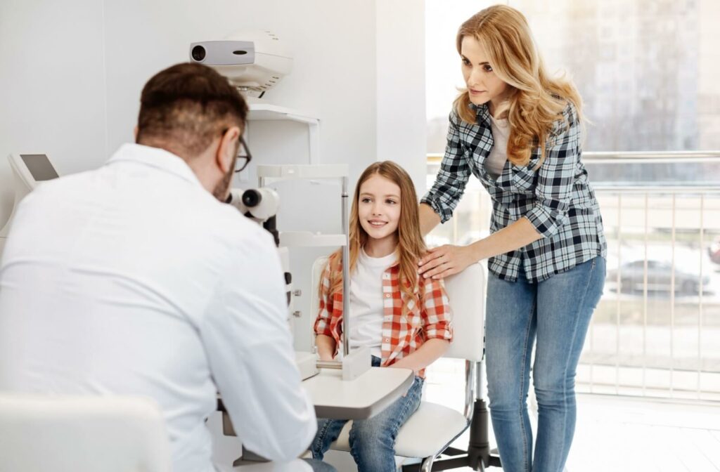 A young girl in a red plaid shirt smiles during an eye exam while her mother stands beside her and an optometrist conducts the assessment