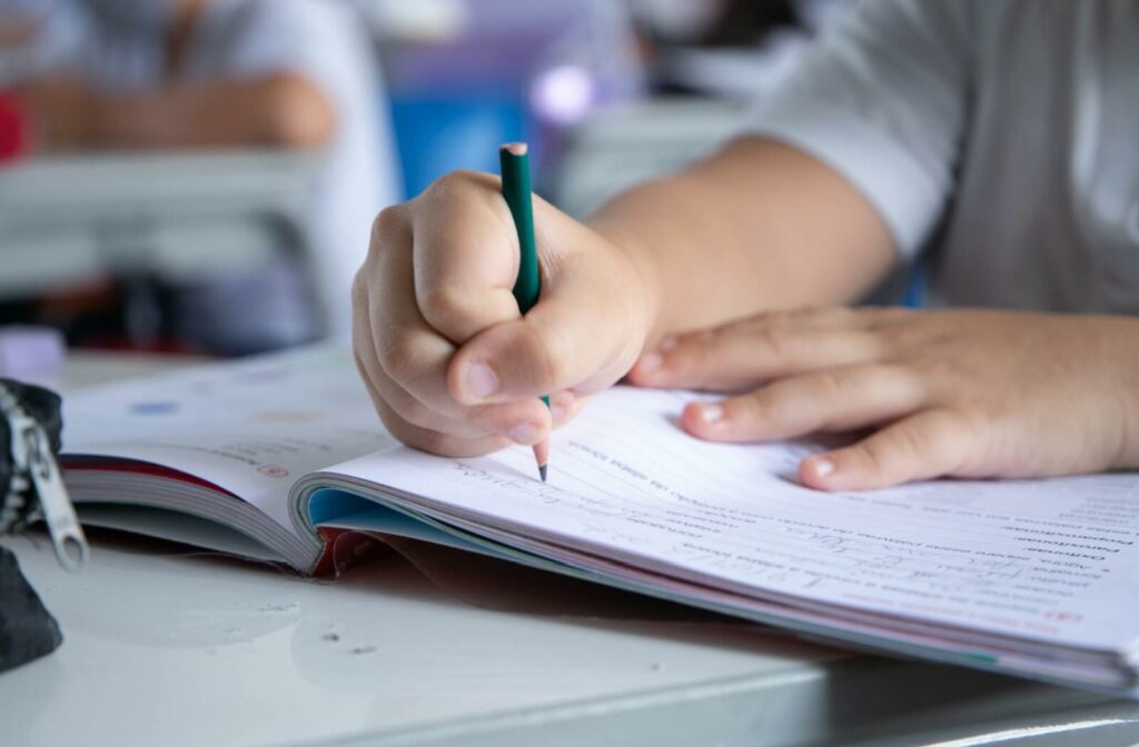 A close-up of a student’s hands writing in a workbook with a pencil during a classroom lesson.