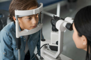 A young boy resting his chin on a slit lamp biomicroscope while an optometrist examines his eye structure during a pediatric myopia control evaluation.