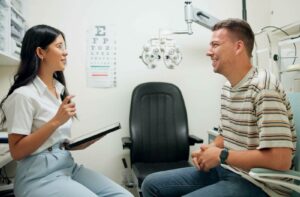 A female optometrist holding a digital tablet while consulting with a smiling adult male patient in an eye examination room with an eye chart in the background.