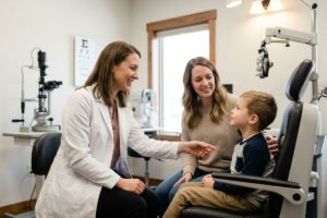 Smiling optometrist reaches toward a young child seated in an exam chair beside an accompanying adult in an eye exam room.